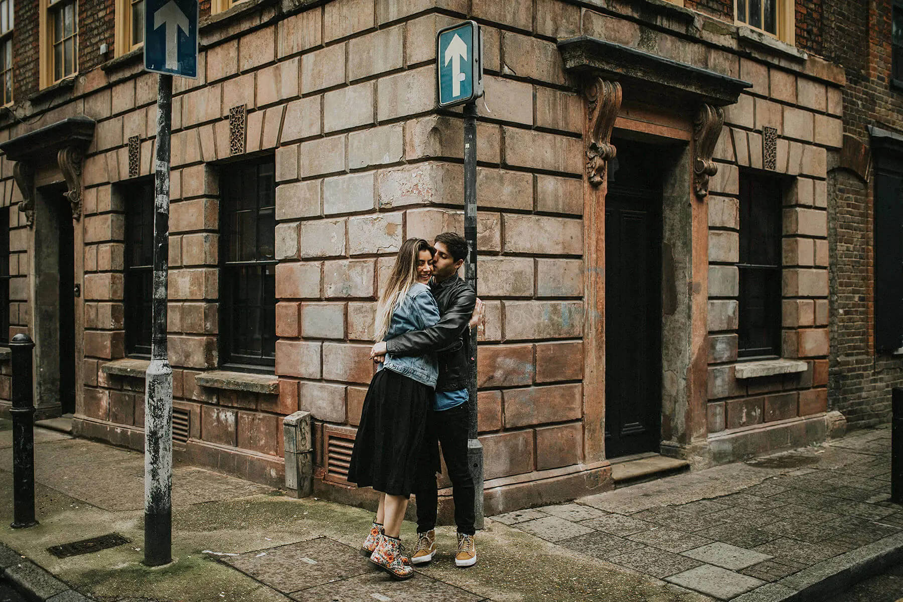  In an old city alley, a couple shares an embrace on the cobblestone sidewalk. The woman, in a black skirt and denim jacket, is smiling as she hugs the man, who is wearing a black leather jacket and jeans. Behind them, a vintage building with brown stone faÃ§ade and barred windows adds a historical touch to the romantic scene. An arrow sign pointing upwards adds a symbolic element to the composition.